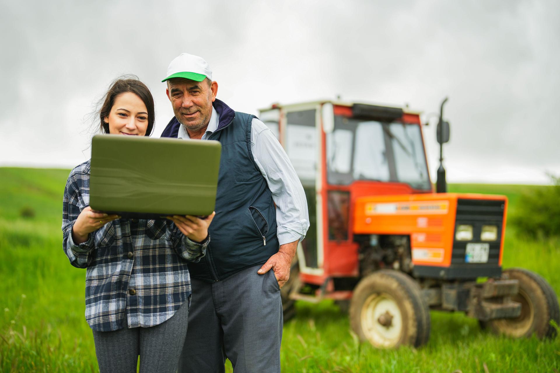 young female agronomist and senior farmer controlling wheat field with laptop with smart farming apps tractor in background